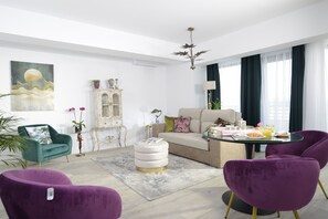 A modern living room with a sofa, round ottoman, and purple chairs. There is a dining table with a lamp and a painting on the wall. - The Clock (Voluntari)