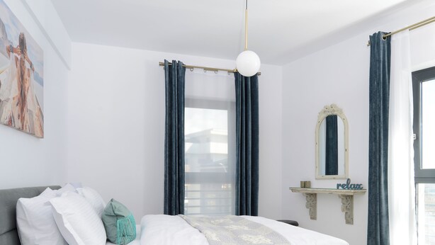 Ein modernes Schlafzimmer mit einem grauen Bett, weißer Bettwäsche, einem türkisfarbenen Zierkissen, einem Glastisch, einer goldfarbenen Lampe und einem Schild mit der Aufschrift 'relax'.