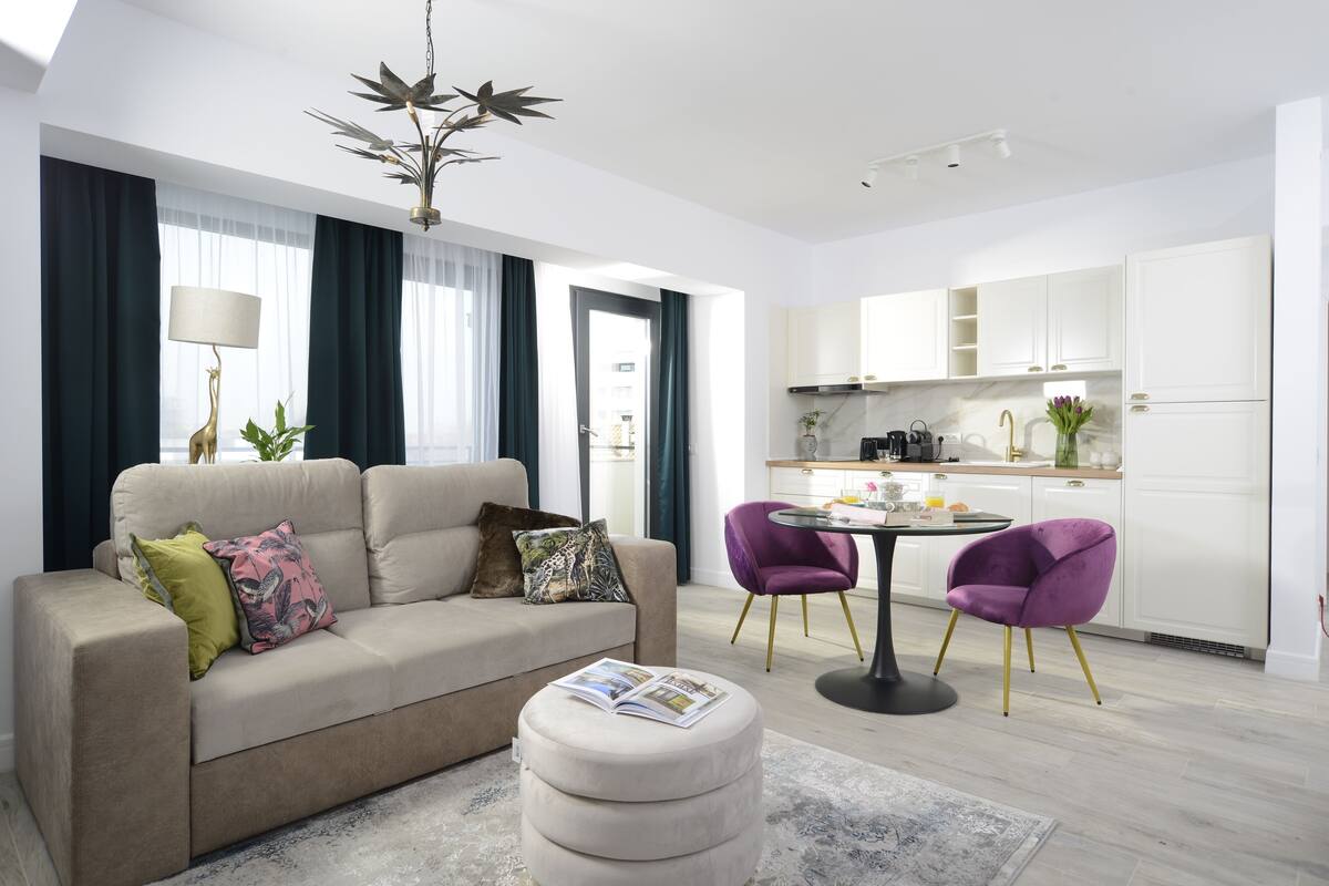 A modern living room with a sofa, ottoman, and two purple chairs. A dining table with a lamp and a kitchen area with cabinets and a sink are visible.