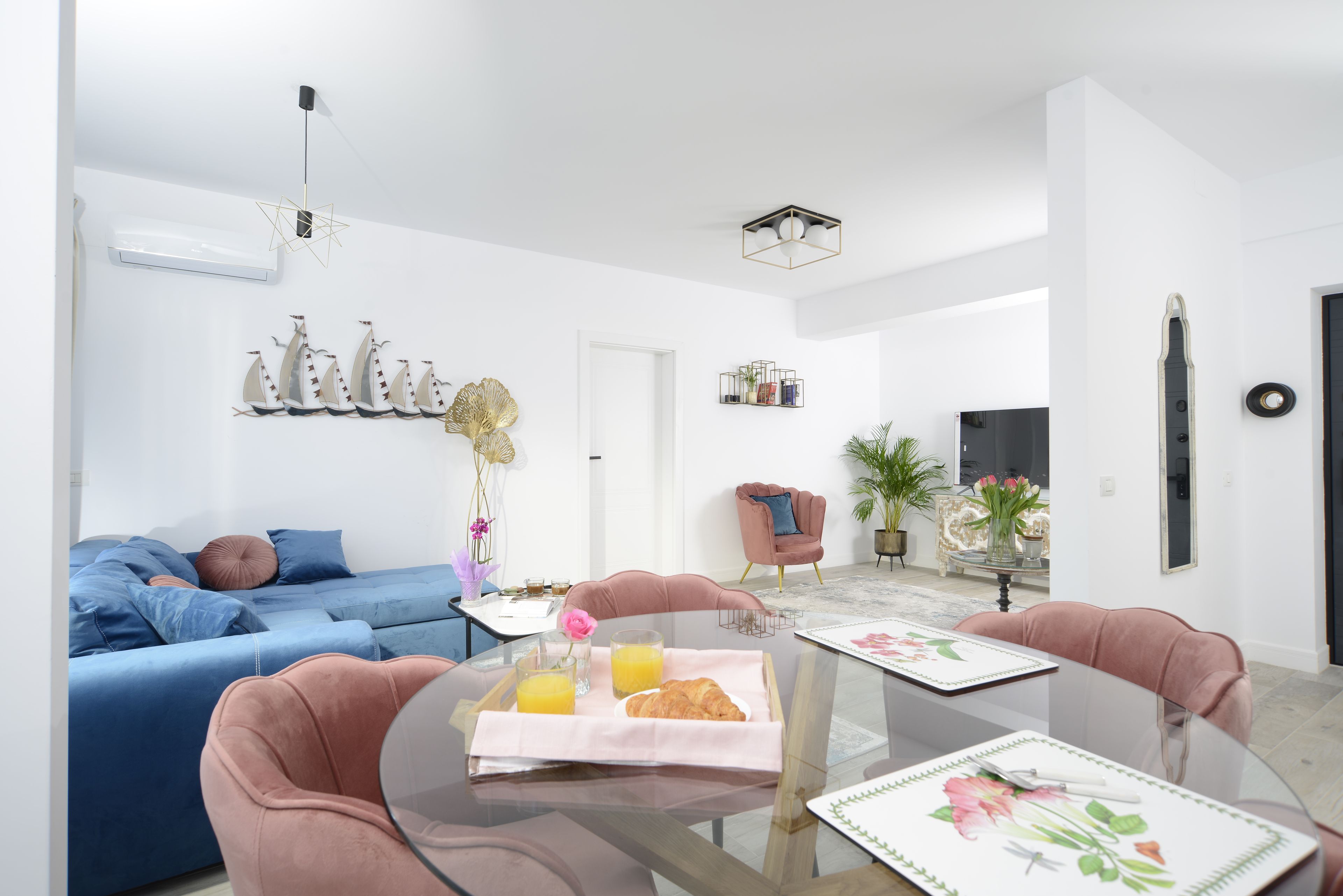 Una sala de estar moderna con un sofá azul, un sillón rosa y una mesa de comedor de vidrio con una bandeja de croissants y jugo de naranja.