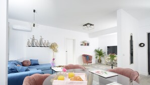 Uma sala de estar moderna com um sofá azul, uma poltrona rosa e uma mesa de jantar de vidro com uma bandeja de croissants e suco de laranja.