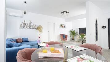 Una sala de estar moderna con un sofá azul, un sillón rosa y una mesa de comedor de vidrio con una bandeja de croissants y jugo de naranja.