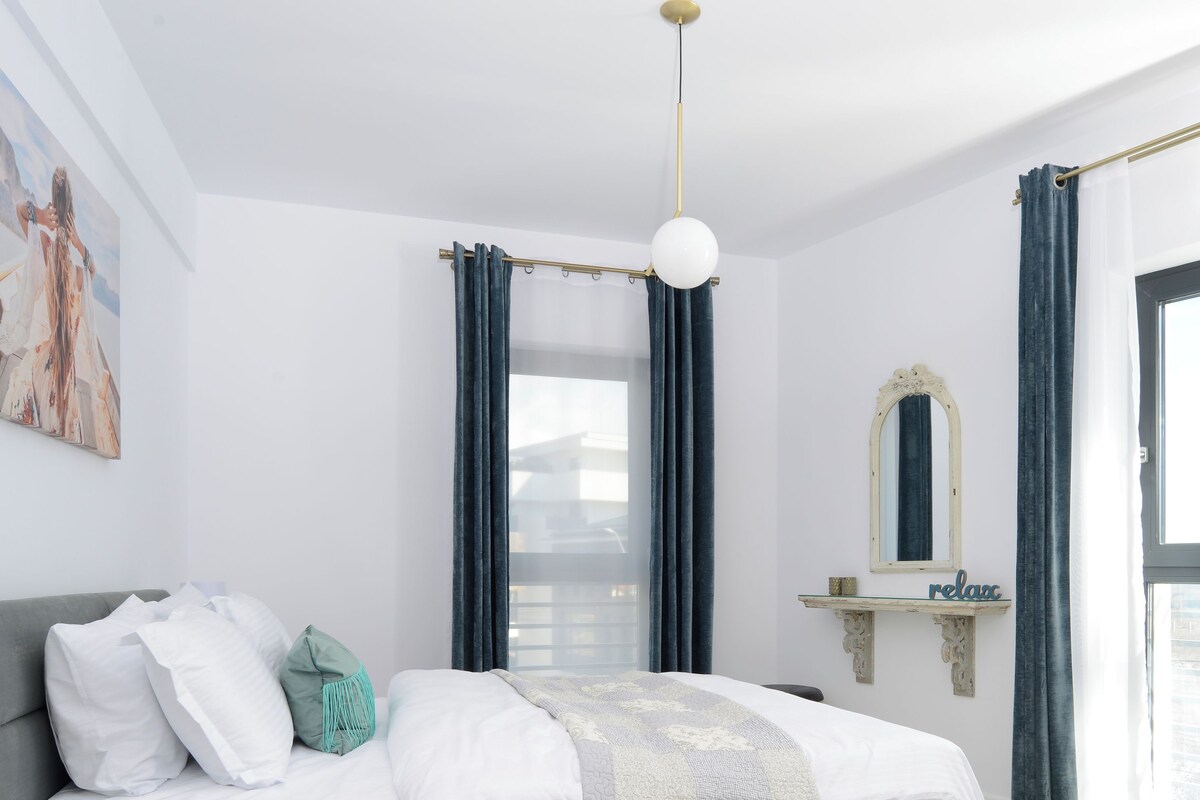 Un dormitorio moderno con cama gris, ropa de cama blanca, un cojín turquesa, una mesita de cristal, una lámpara dorada y un cartel con la palabra 'relax'.