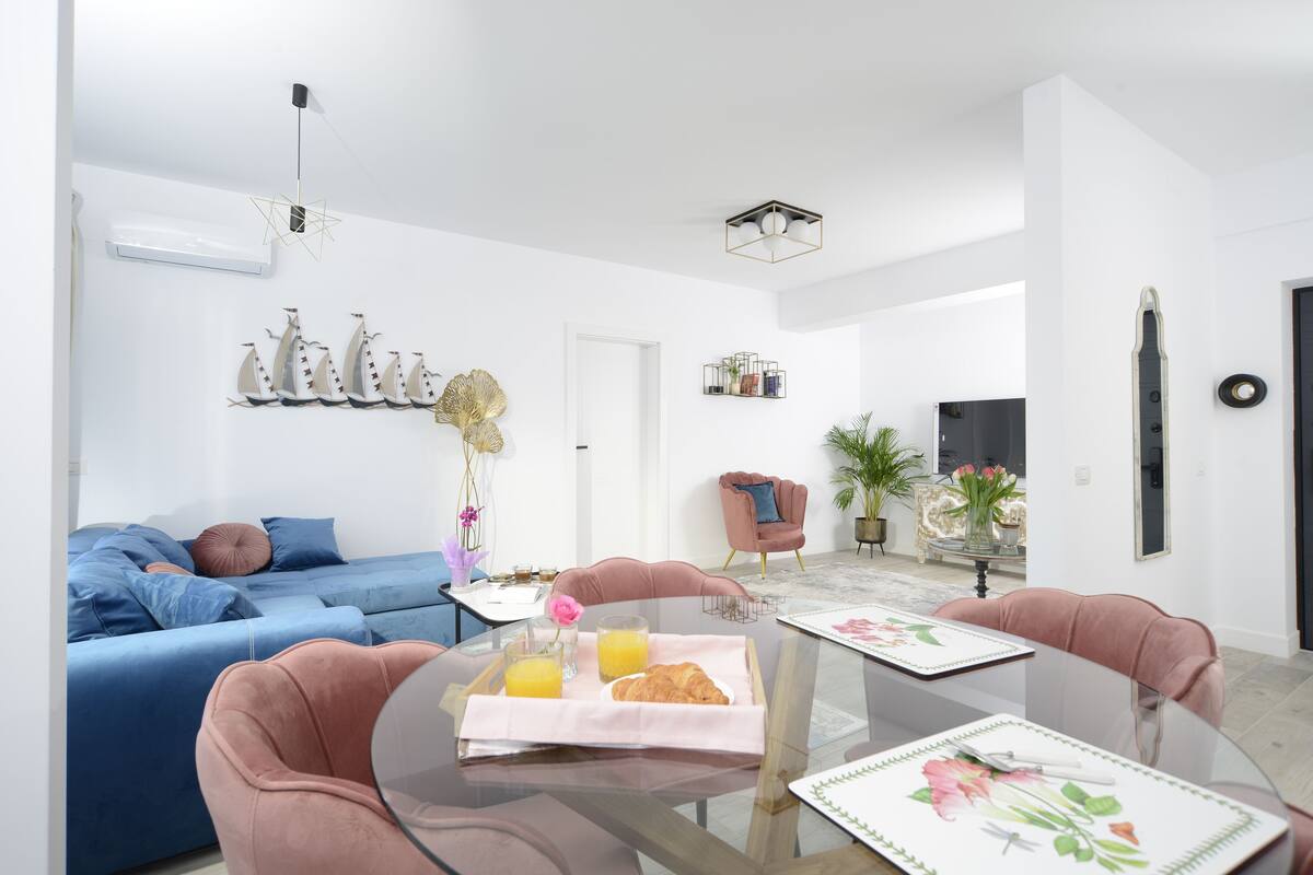 A modern living room with a blue sofa, a pink armchair, and a glass dining table with a tray of croissants and orange juice.