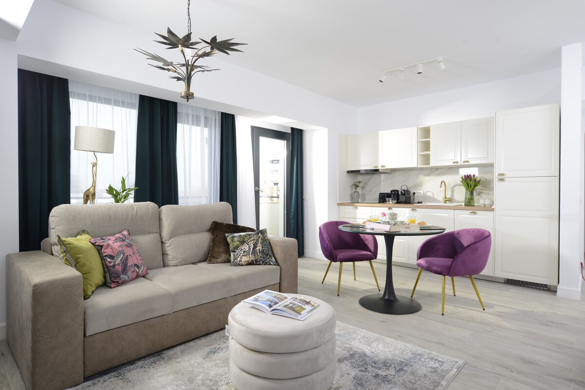 A modern living room with a sofa, ottoman, and two purple chairs. A dining table with a lamp and a kitchen area with cabinets and a sink are visible.