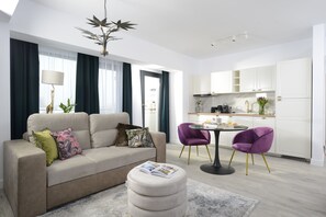 Un salon moderne avec un canapé, un pouf et deux fauteuils violets. On aperçoit une table à manger avec une lampe, ainsi qu’un coin cuisine doté de placards et d’un évier.