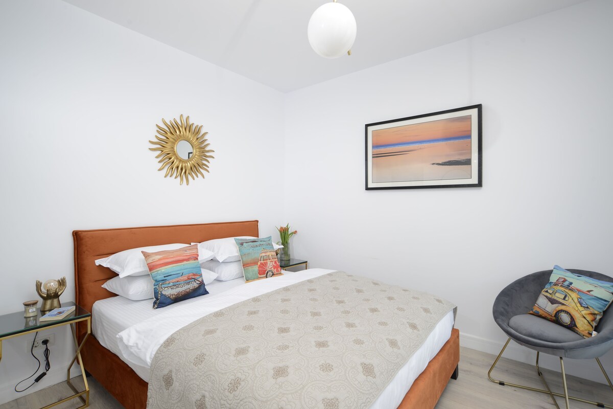 A neatly arranged bedroom with a bed, a bedside table, a chair, a wall-mounted mirror, and a framed picture.