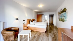 A modern hotel room with a wooden bed, a white table, a brown armchair, and a painting on the wall. - Comfortable and versatile room -perfect for relaxation, work and exchange (Winterberg)