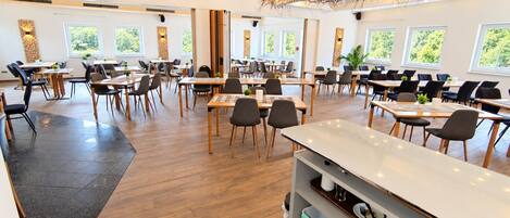 A modern dining area with multiple tables and chairs, large windows, and a unique ceiling design.