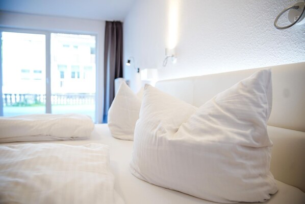 A hotel room with a bed, pillows, a nightstand, a lamp, and a view of the outside.