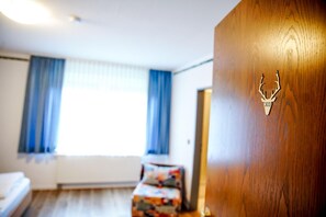 Et hotelværelse med en seng, en trædør med et skjoldmærke af et rådyr, og et vindue med gardiner.