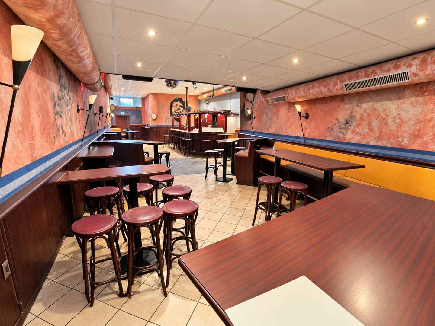 A restaurant interior with multiple wooden tables and red stools, a bar area, and a tiled floor.