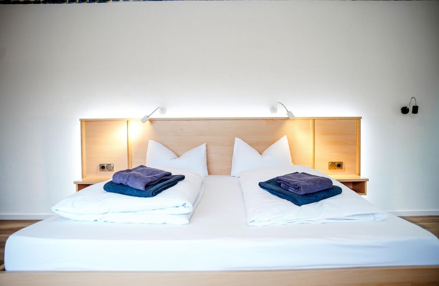Uma cama de casal com roupa de cama branca, duas mesas de cabeceira com abajures e uma cabeceira de madeira.