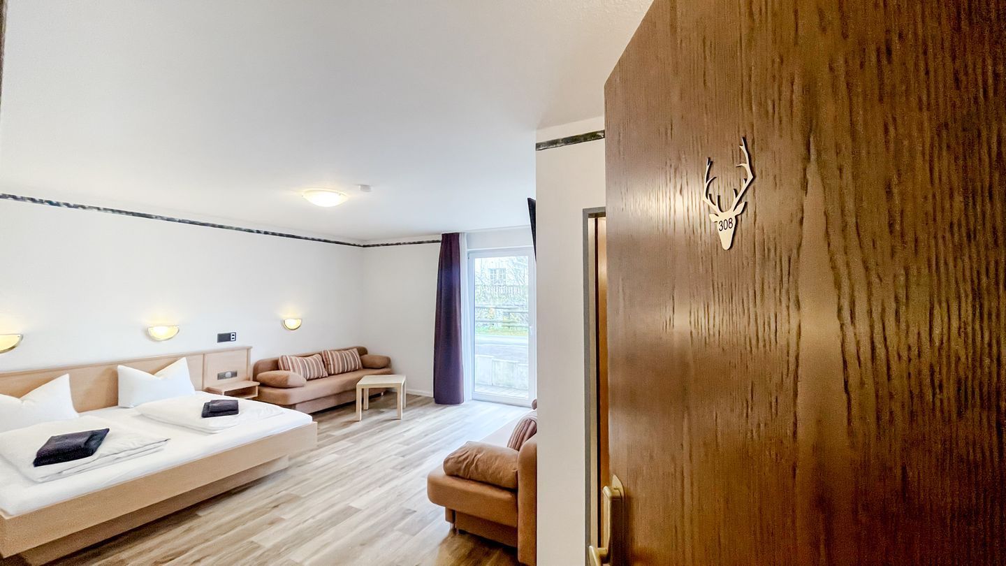 Ett modernt hotellrum med en säng, två soffor, ett litet bord och en trädörr prydd med en hjorthuvudsymbol.