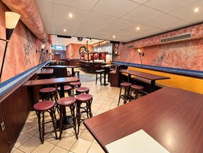 Interior de restaurante con varias mesas de madera y taburetes rojos, una zona de bar y piso de baldosas.