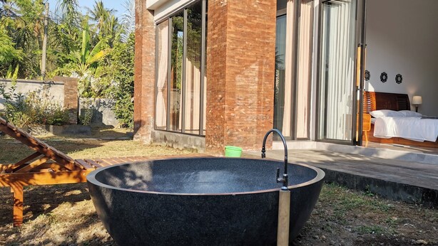 A modern outdoor bathroom with a large black bathtub, a wooden deck, and a view of a bedroom with a bed and curtains. - Villa Kiprey (Karangasem)