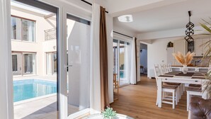 Un salon moderne doté d’une grande porte vitrée donnant sur une piscine, d’un coin repas avec une table et des chaises blanches, et d’une horloge posée sur le sol.