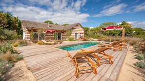 Une terrasse en bois avec une piscine, des chaises longues et un bâtiment dont le toit est recouvert de chaume.