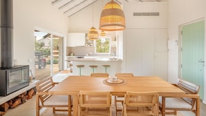 Une cuisine moderne avec des meubles blancs, une grande table à manger en bois, des chaises en osier et des suspensions en osier.
