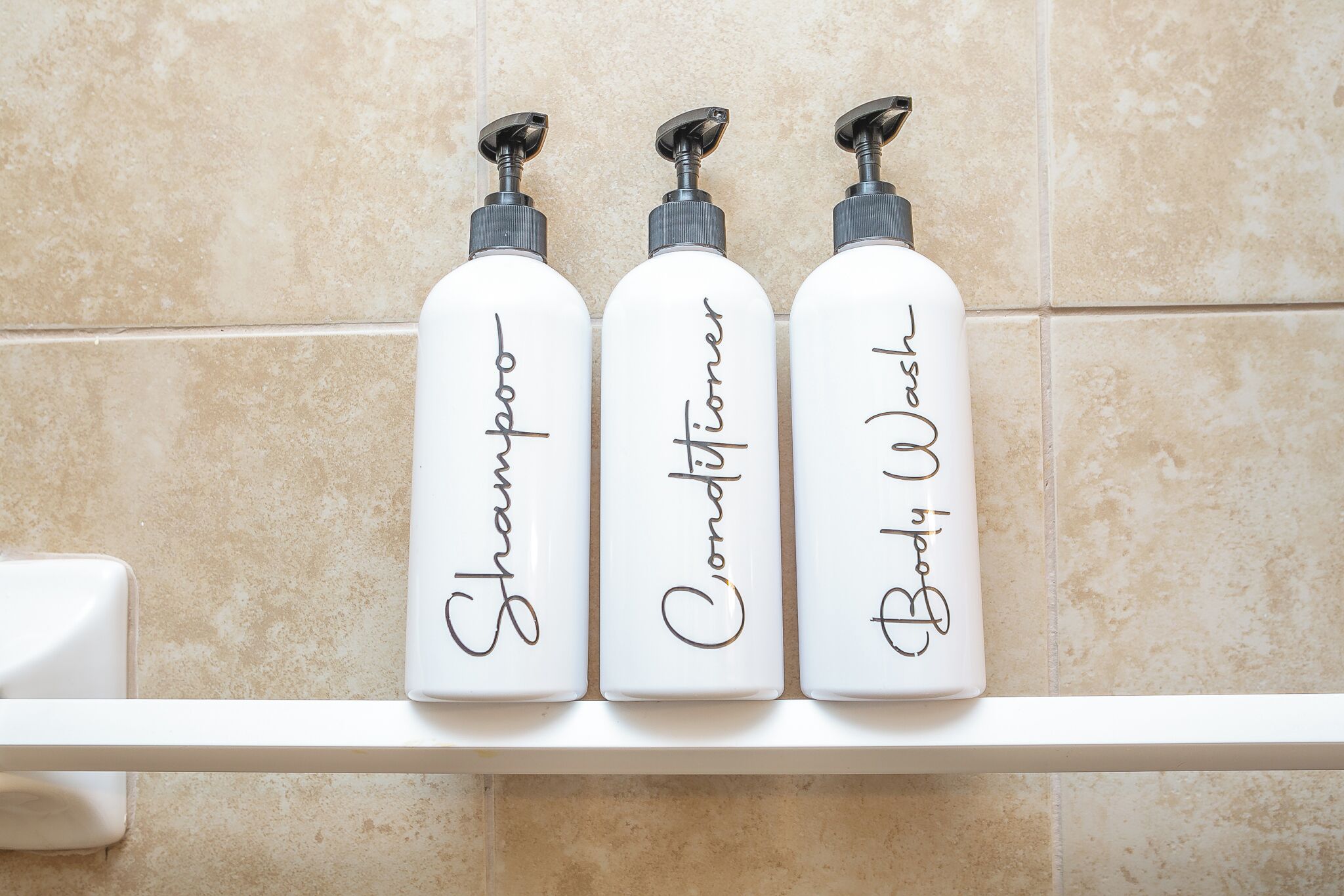 Trois bouteilles de shampoing, d’après-shampoing et de gel douche, posées sur une étagère blanche devant un mur carrelé.