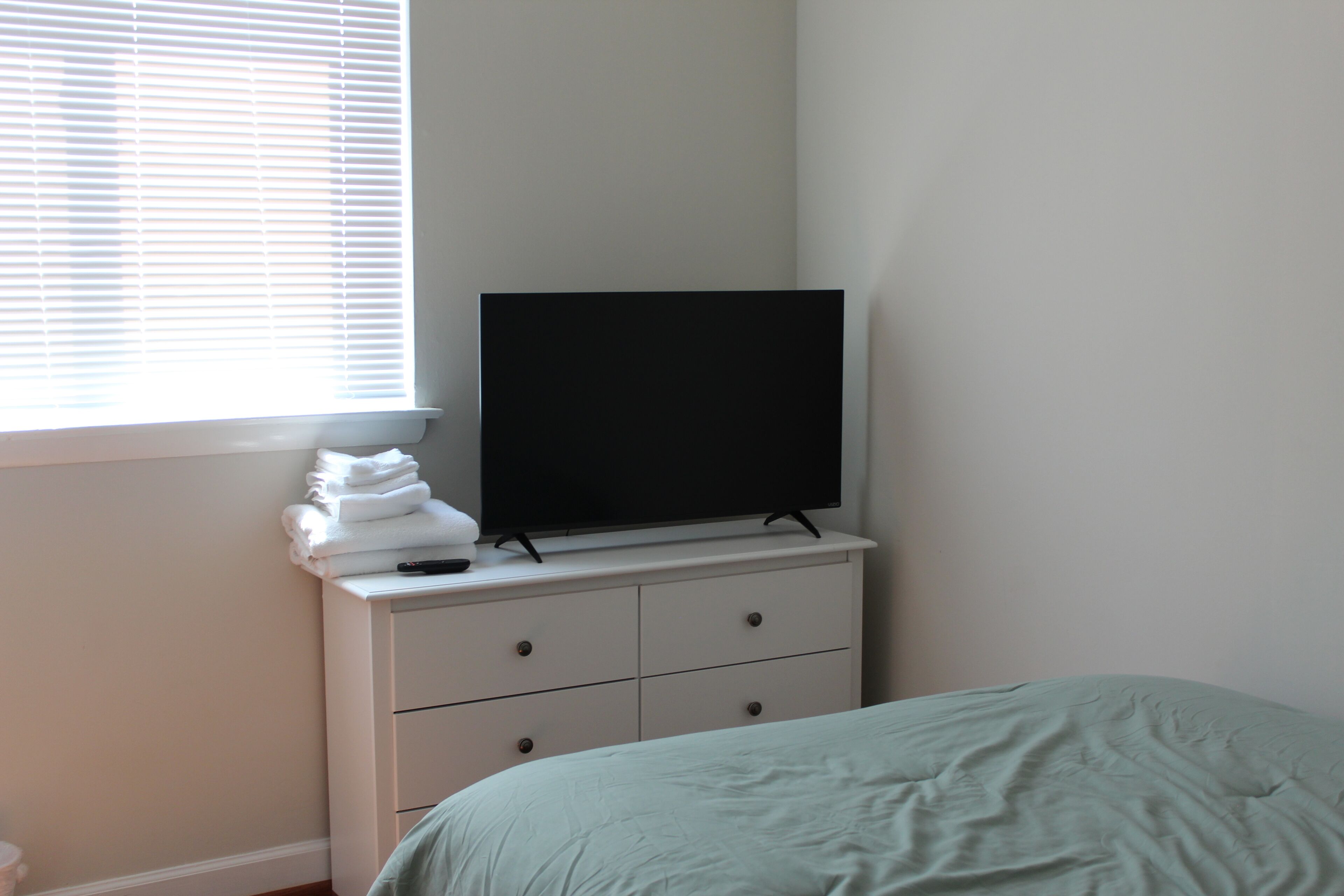 Ein Schlafzimmer mit einem Bett, einer Kommode mit Fernseher darauf und einem Fenster mit Jalousien.