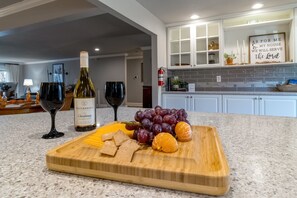Una cocina con encimera de mármol, tabla de cortar con uvas, naranjas y queso, y una botella de vino.