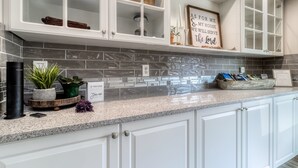 Une cuisine avec des meubles blancs, un plan de travail en granit et un pare-feu recouvert de carreaux gris.
