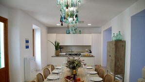 A dining area with a long table set for six, surrounded by wicker chairs. A decorative chandelier hangs above, and a potted plant is on the kitchen counter.