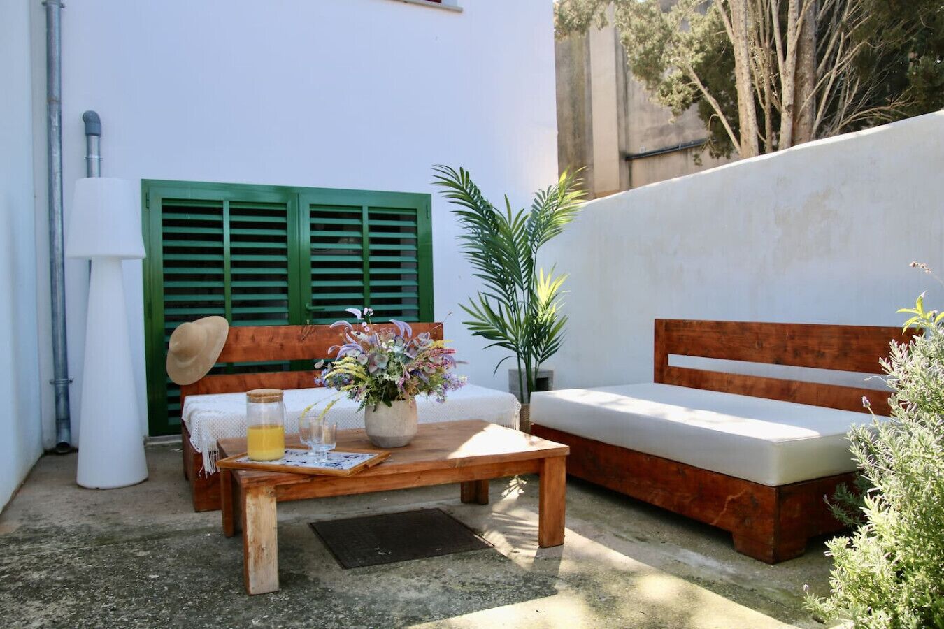 Un espace extérieur aménagé avec un banc et une table en bois, un pot de fleurs et une porte verte à volets.