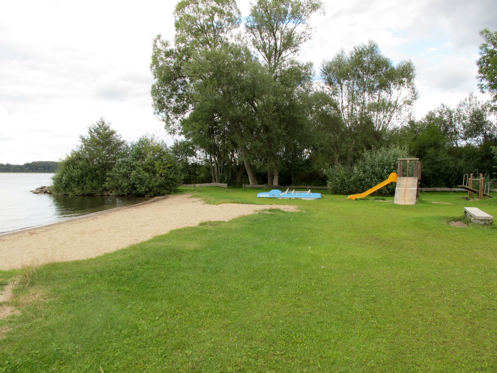 Zona verde con un parque infantil, que incluye un tobogán y columpios, cerca de un cuerpo de agua.