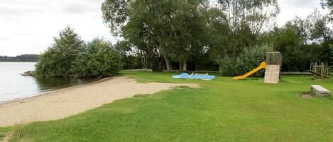Zona verde con un parque infantil, que incluye un tobogán y columpios, cerca de un cuerpo de agua.