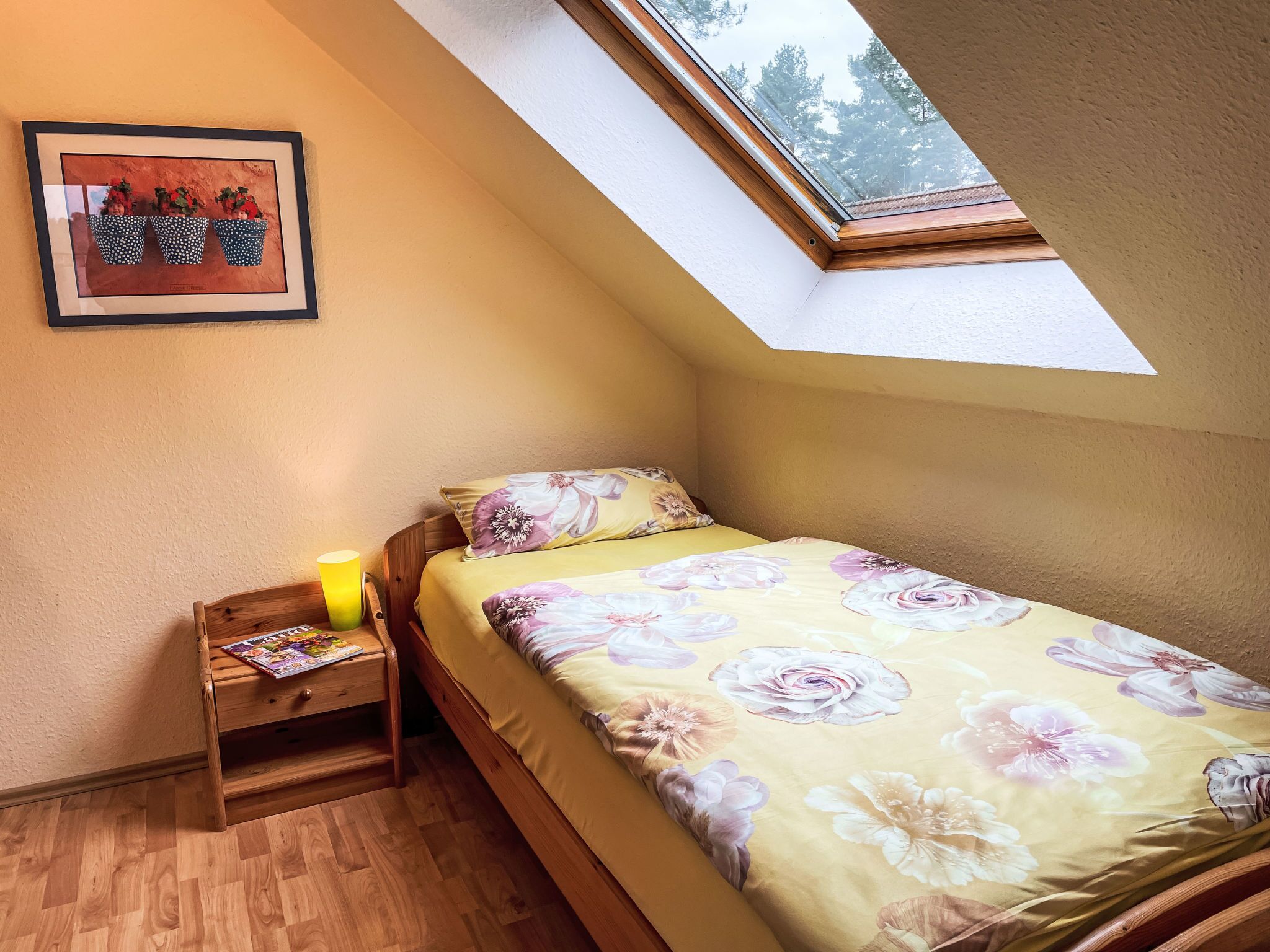 A bedroom with a sloped ceiling, a wooden nightstand with a lamp, a framed picture, and a bed with floral bedding.
