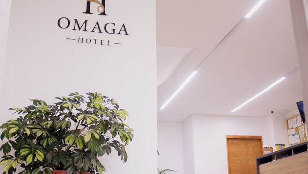 Ett entréområde med en krukväxt, en receptionsdisk och en vägg med hotellets namn, ”OMAGA HOTEL”.