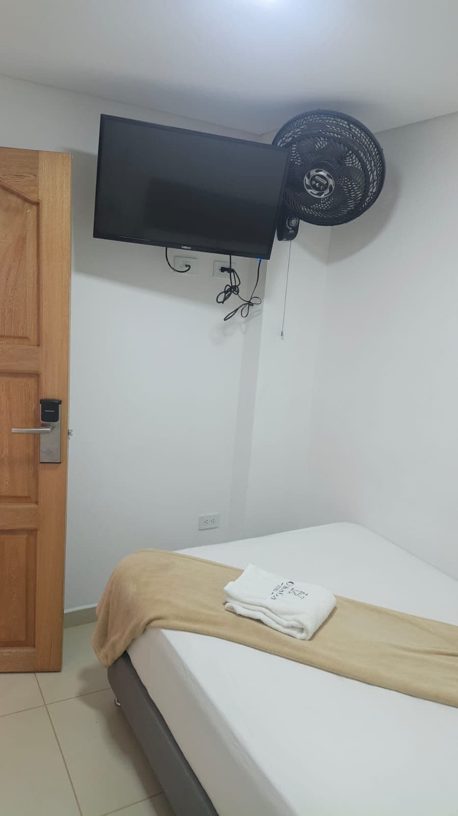 Una habitación con una cama, un televisor de pantalla plana montado en la pared, un ventilador de techo y una puerta de madera.