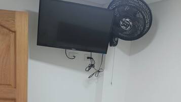 Una habitación con una cama, un televisor de pantalla plana montado en la pared, un ventilador de techo y una puerta de madera.