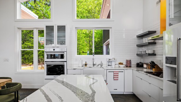 A modern kitchen with white cabinetry, a central island, and large windows with a view of trees. - Fall Mtn Views | Hot Tub · Game Room · 10m AVL (Asheville)
