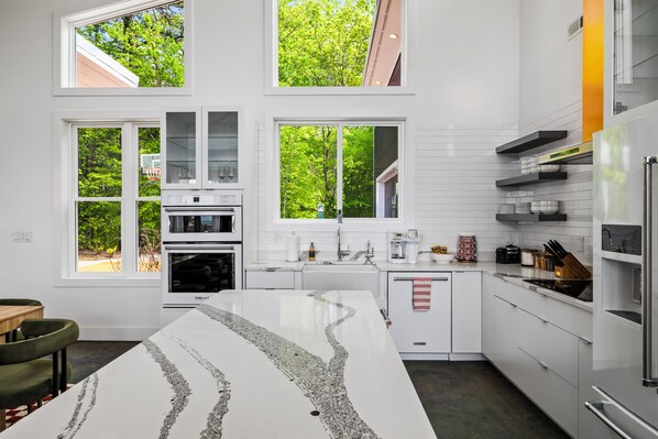 Una cocina moderna con armarios blancos, una isla central y grandes ventanales con vistas a árboles.