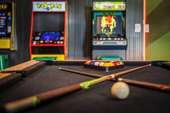 A pool table with balls and cues, surrounded by arcade machines.
