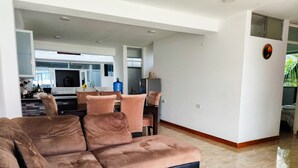 A spacious living room with a large brown sectional sofa, a dining area with a pink chair, and a kitchen in the background.