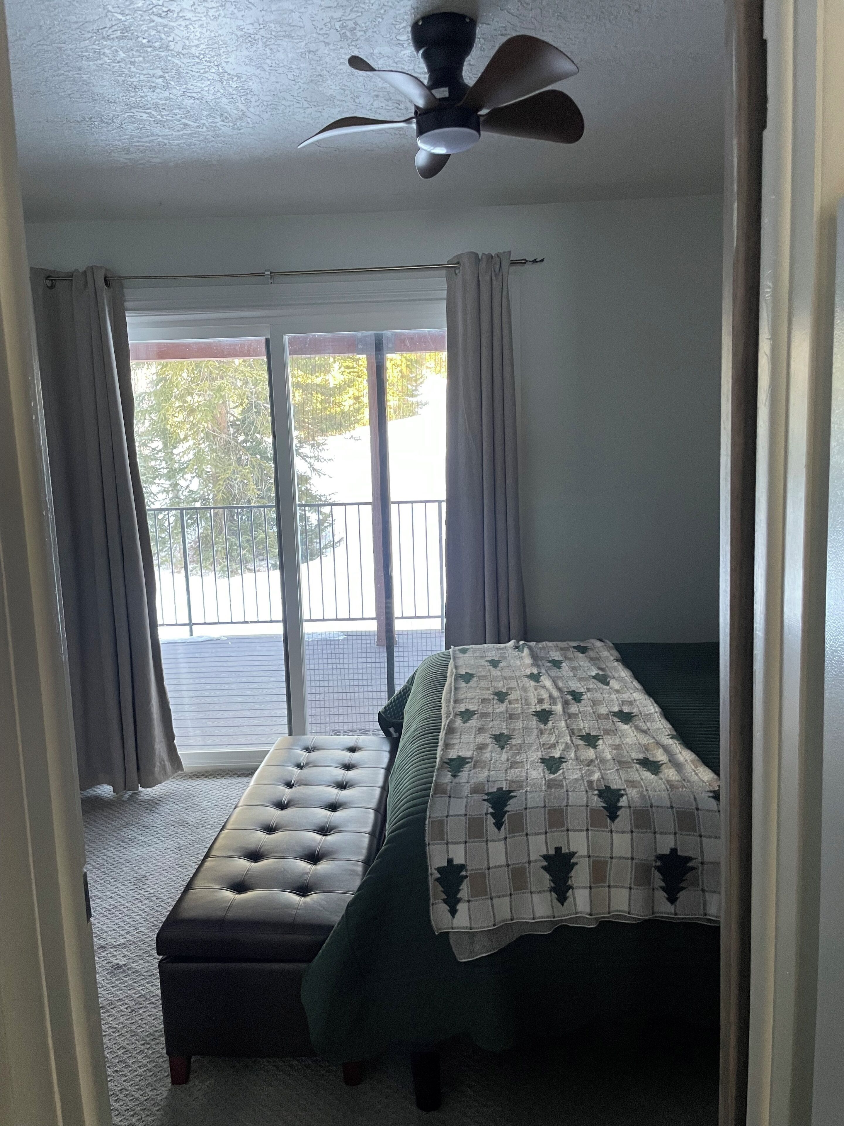 A bedroom with a bed, a bench, a ceiling fan, and a view of the outdoors through a sliding door.