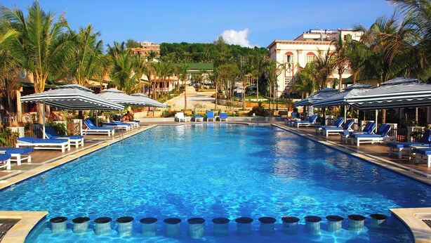 A swimming pool with lounge chairs and umbrellas, surrounded by palm trees and buildings. - PV BEACH BUNGALOW (Phu Quoc)