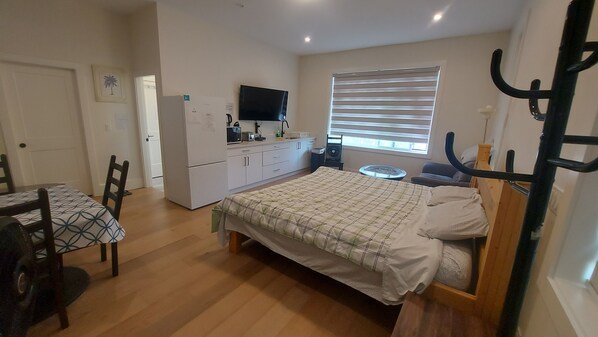 A compact bedroom with a bed, a small table, a chair, a kitchenette, and a window with blinds. - Self Contained Master Suite (Surrey)