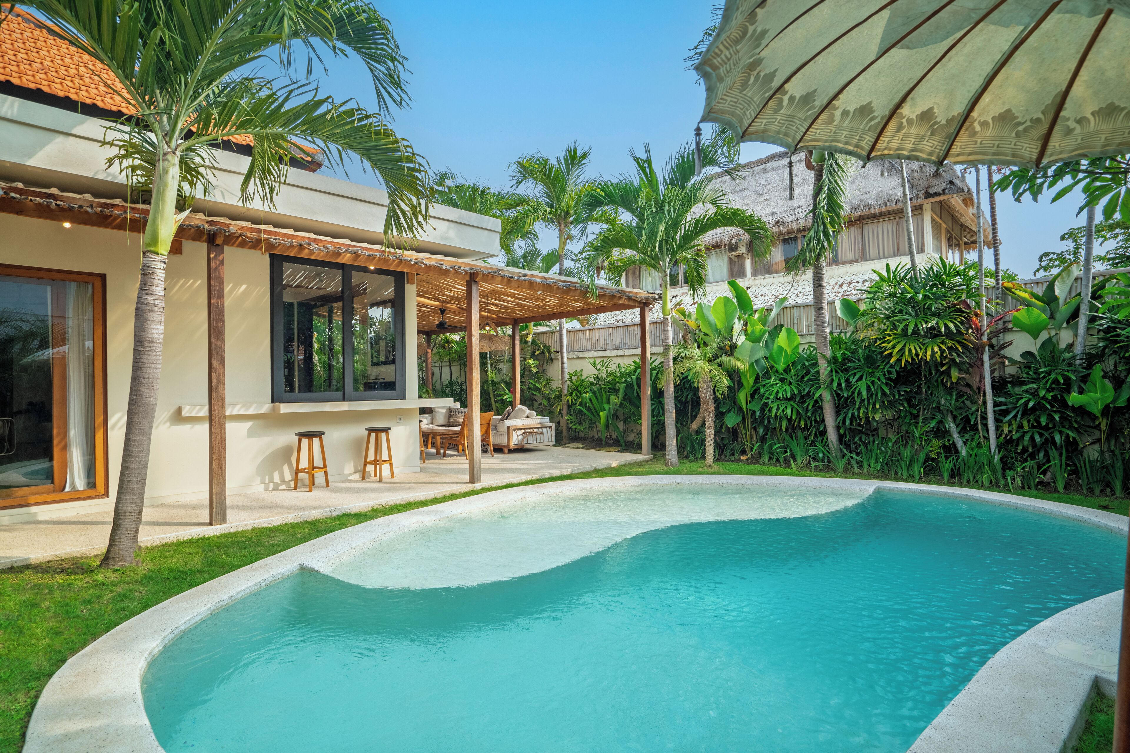 A swimming pool with a sunken seating area, surrounded by lush greenery and a covered outdoor seating area.