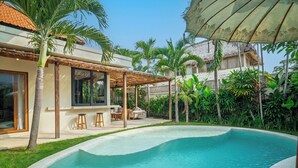 Una piscina con zona de estar hundida, rodeada de exuberante vegetación y un área de estar techada al aire libre.