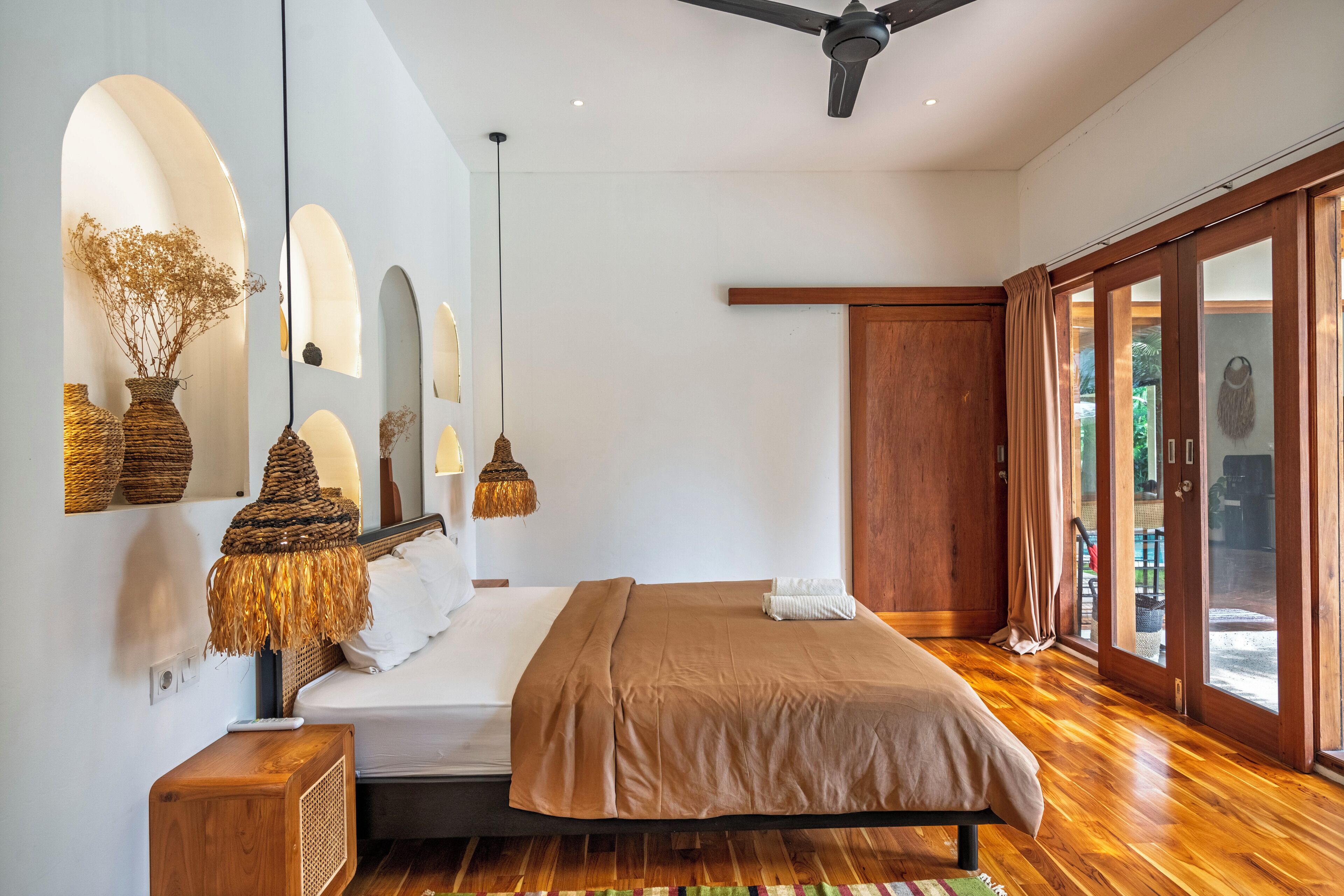 Un dormitorio moderno con piso de madera, una cama con colcha marrón, una mesita de noche con lámpara y vistas al exterior por una puerta corredera.
