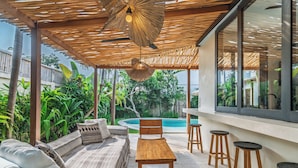 Un espacio de estar al aire libre con techo, piscina, mobiliario de mimbre y taburetes de madera.