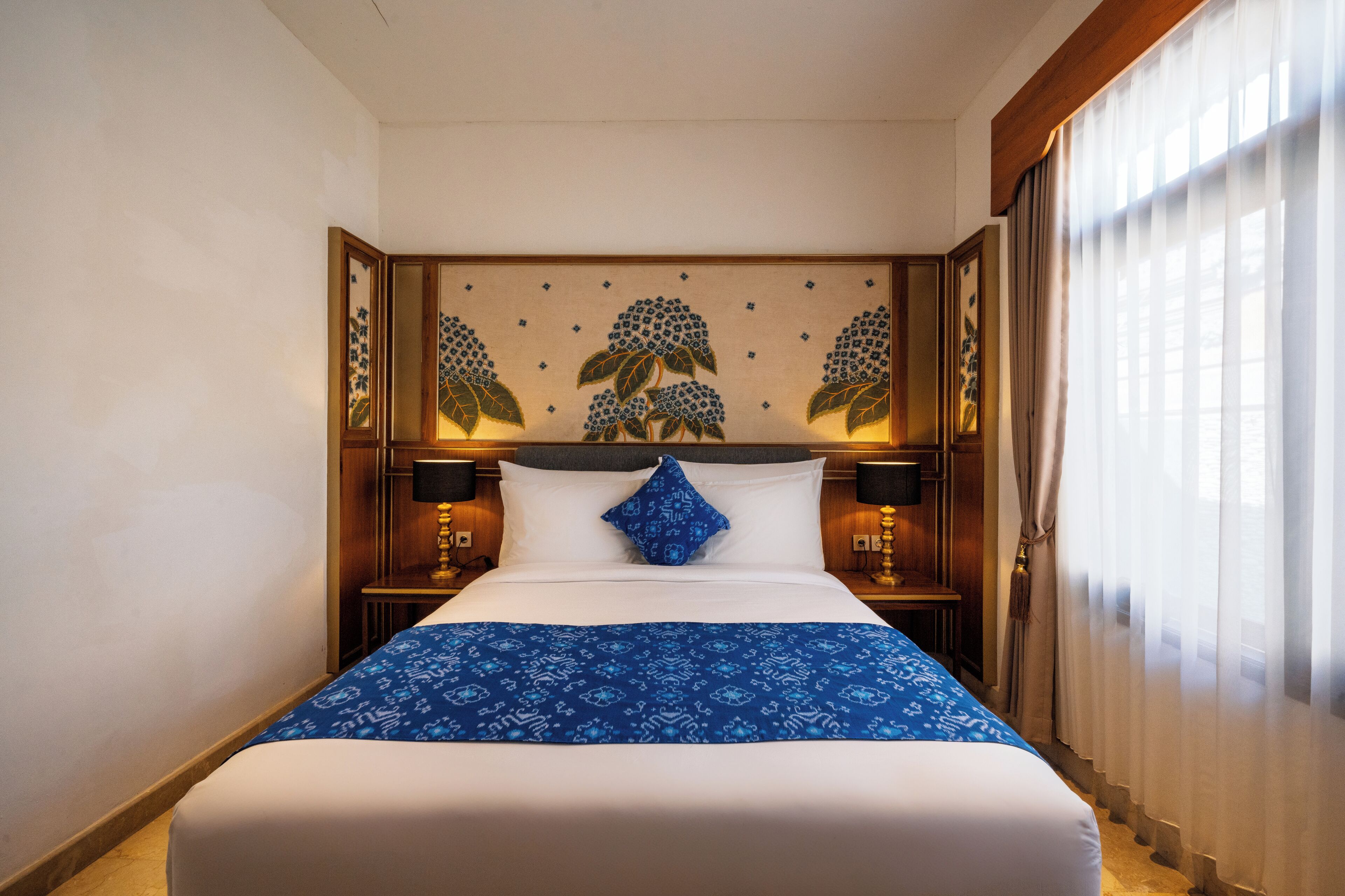 Habitación de hotel con una cama grande, un cabecero decorativo, dos lámparas de noche y una ventana con cortinas translúcidas.