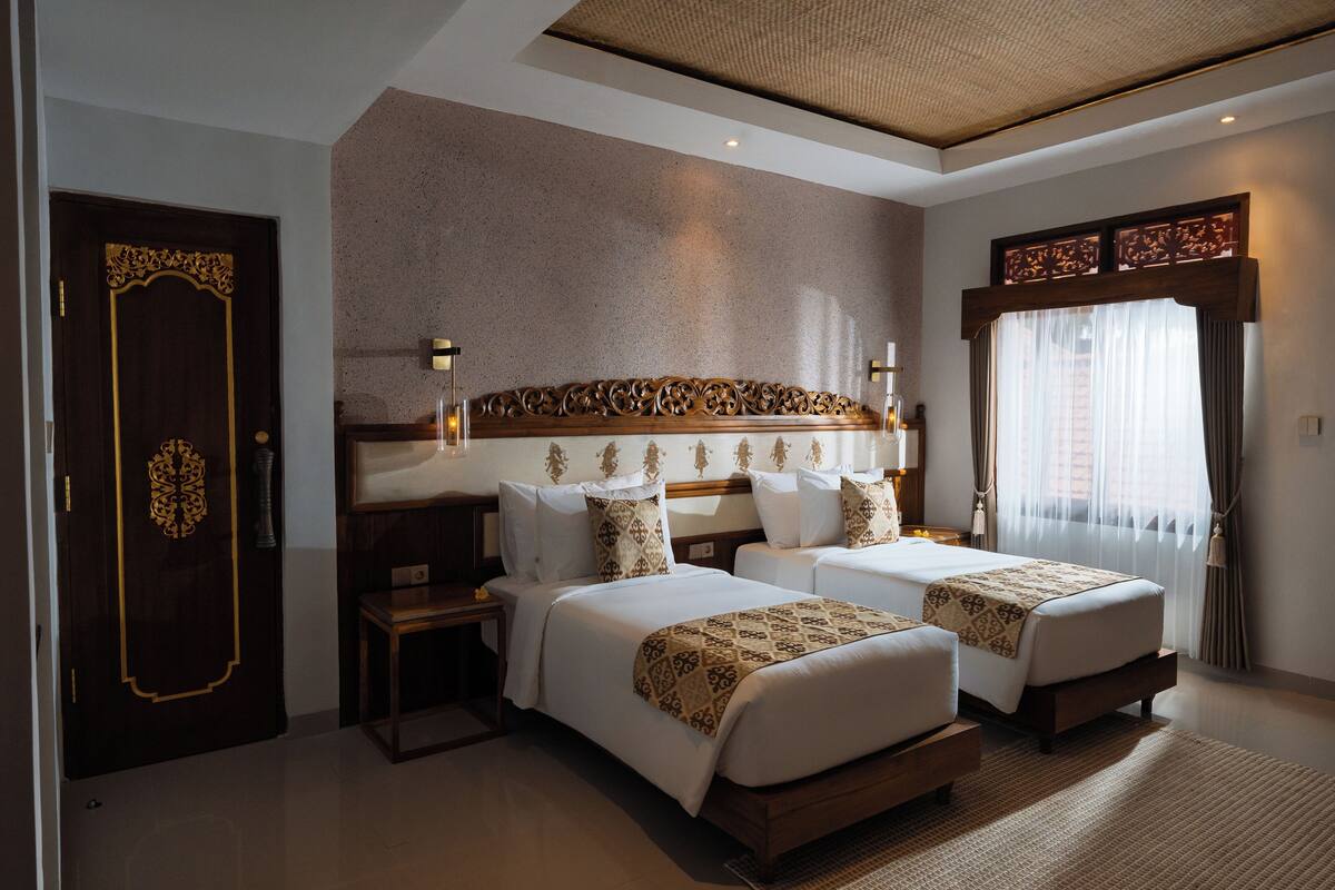 A hotel room with two beds, a large decorative headboard, a wooden door with intricate designs, and a large window with curtains.