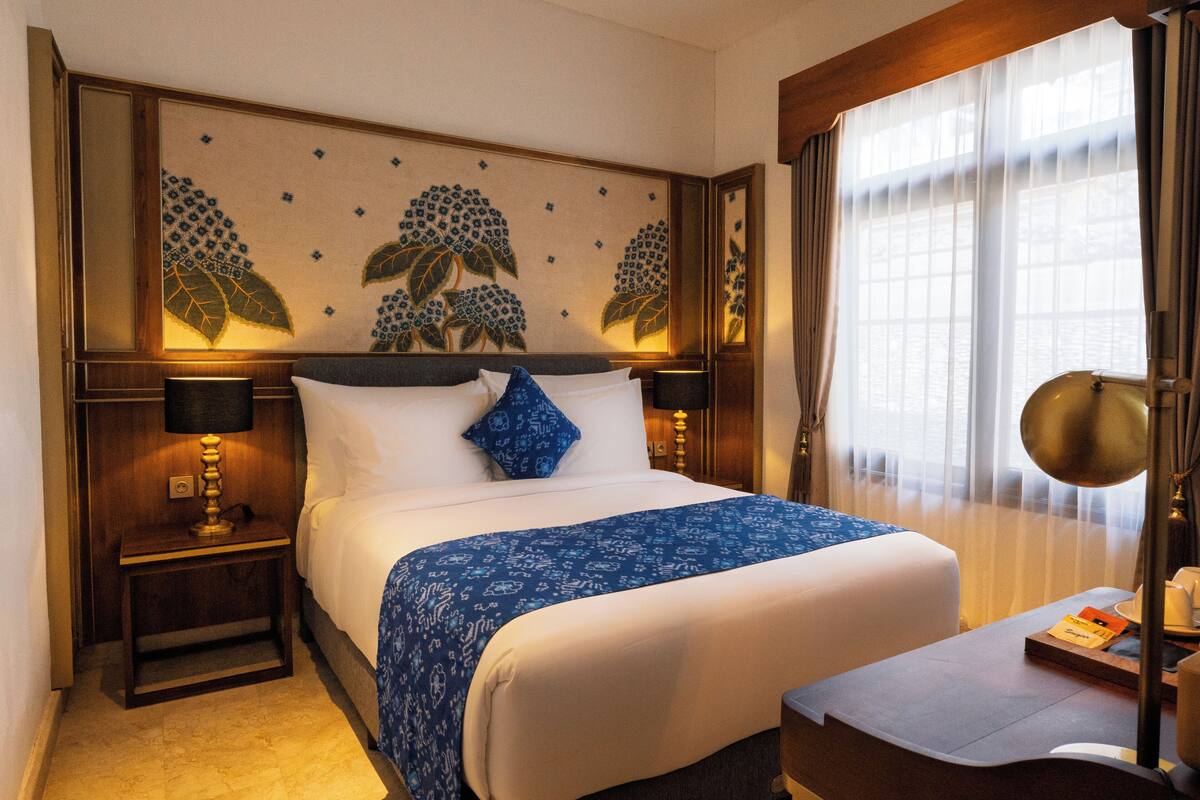 A hotel room with a large bed, a nightstand with a lamp, a bedside table with a lamp, and a view of the outdoors through sheer curtains.