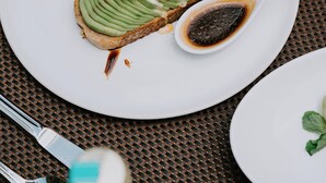 A white plate with a sandwich topped with avocado slices, a poached egg, and a side salad, accompanied by a white spoon with sauce. - Puri Kantor Legacy (Ubud)